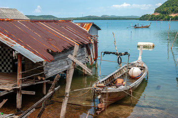 Thai fishing long boat next to the fisherman's hut standing on stilts in the water. Rusty Shack Roof. A fisherman pours water from a boat. Big buoy in the boat.