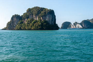 A large rock in the turquoise sea and a white sailing boat nearby. The rock is all overgrown with trees. The sky without clouds.