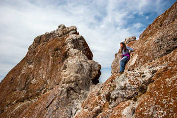 A girl is standing on a rock, cuddling her back and resting her arms. European appearance, long black hair, pants. Clouds in the sky. The rock is covered with red moss.