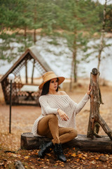 Portrait of beautiful brunette woman sitting near the lake, wooden arbor and stairway. Fashion outdoor shot of pleased lady in cozy clothes and straw hat enjoying nature views in autumn countryside.