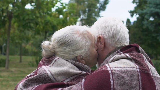 Elder couple sitting on bench under plaid, mature woman kissing man on cheek