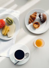 Assorted breakfast with a cup coffee, fruits, croissant and cupcake on white plate. Breakfast at hotel on white table with direct light coming from the window.