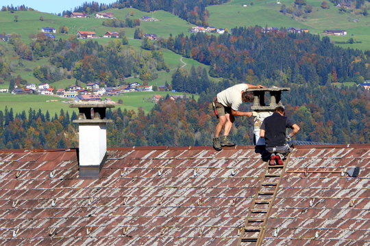 Renovierungsarbeiten an einem Kamin, zwei Handwerker stehen auf einem Dach und reparieren einen Kamin