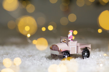 Decorative figurines of a Christmas theme. Santa statuette rides on a toy car with a trailer for gifts. Festive decor, warm bokeh lights.