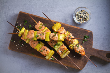 Skewered salmon teriyaki with leeks, sprinkled with sesame seeds on serving board. Overhead, horizontal