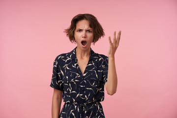 Studio shot of angry brunette female with casual hairstyle looking at camera with wide mouth opened and frowning face, posing over pink background with raised hand