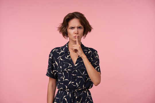 Serious young pretty woman with short brown hair keeping raised forefinger on her lips, looking at camera and frowing eyebrows while standing over pink background