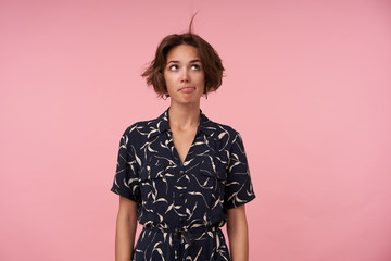 Attractive young brunette lady with short haircut wearing patterned elegant dress, posing over pink...