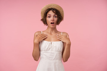 Surprised pretty brunette woman with casual hairstyle wearing white summer elegant dress with straps, holding palms on her chest and looking to camera with round eyes, standing over pink background