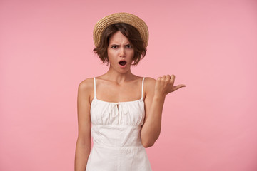Portrait of young brunette female in white summer looking at camera with confused face, showing aside with thumb while standing over pink background, frowning her eyebrows with opened mouth