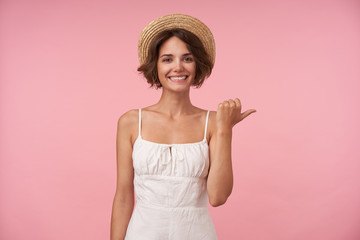Cheerful pretty young brunette woman with casual hairstyle pointing aside with raised thumb while looking happily to camera, isolated over pink background in white elegant dress and boater hat