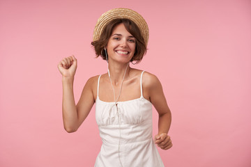 Joyful young brunette woman with short haircut looking at camera with wide smile and wearing earphones, enjoying fine music and dancing over pink background