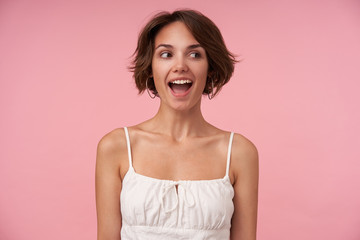 Joyful attractive brunette female with short haircut looking aside with wide happy smile, keeping hands along her body, posing over pink background, being in high spirit