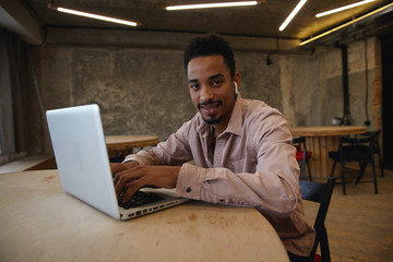 Young handsome bearded freelancer with dark skin working remotely with modern laptop in coworking space, keeping hands on keyboard and looking to camera cheerfully