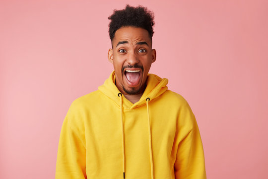 Indoor photo of young bearded dark skinned man looking at camera with wide eyes and mouth opened and wrinkling his forehead, wearing yellow sweatshirt while posing over pink background