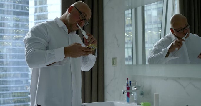 busy bald man is cleansing teeth by electric toothbrush and looking on screen of smartphone