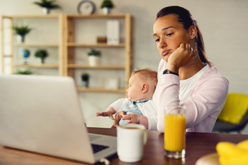 Young pensive mother with baby using laptop at home.