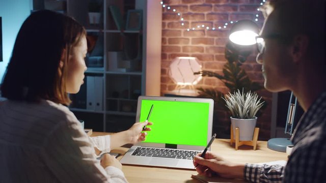 Specialists girl and guy are pointing at green chroma key laptop screen in dark office talking discussing content. People, modern technology and job concept.