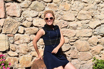 Blond girl wearing sunglasses and silver necklace posing smiling. Medieval stone wall at the background. The French touristic mountain town of Villefranche-de-Conflent with its cobblestone streets. 