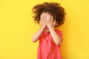 Portrait of little African-American girl hiding her face against color background