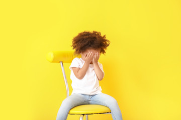 Portrait of little African-American girl sitting on chair against color background