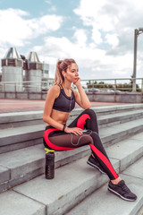 Girl athlete sits on stairs, rest after workout, summer day city. In hand, smartphone listens music headphones. Enjoys listens to podcast lecture. Active lifestyle fitness, fashionable stylish woman.