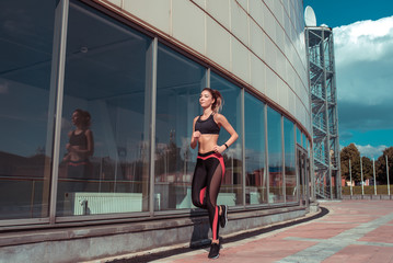 Fototapeta na wymiar Young athletic woman running in summer on jogging in city, background glass windows of building, sportswear. Athletics training, day morning. Active fitness lifestyle, free space text motivation.