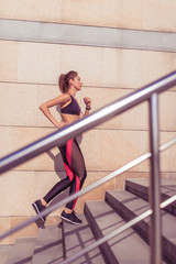 Young and athletic woman, runs up stairs, in summer on jogging in city, side view, sportswear. Workout athletics, day and morning. Active fitness lifestyle.
