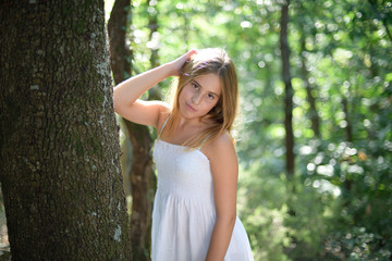 Naklejka premium Girl leaning on a tree in a forest in a white dress and looking