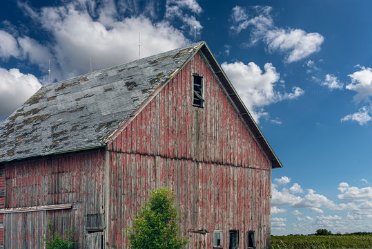 An old, red wooden barn with peeling paint, and shingles coming off the roof.  Lightning rods on top of the roof against a blue sky & fluffy clouds
