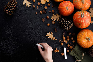 Autumn frame of pumpkins, tree leaves, hazelnuts on a black textural background. Hand with crayons for drawing. Autumn, fall, thanksgiving concept. Flat lay, copy space.