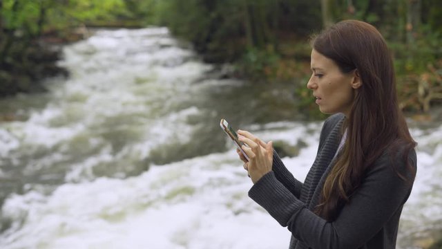 Happy content young attractive woman exploring the country by a fast flowing river with rapids on her social media and cell phone outside during the day