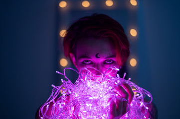 Woman with witch makeup and an earring in her nose. The girl holds a garland of flickering pink...