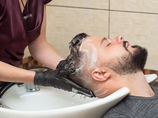 Female washing clients hair and making head massage, close up view. Stylists hands in black rubber gloves. Photographed in hairdressing saloon. Selective soft focus. Blurred background