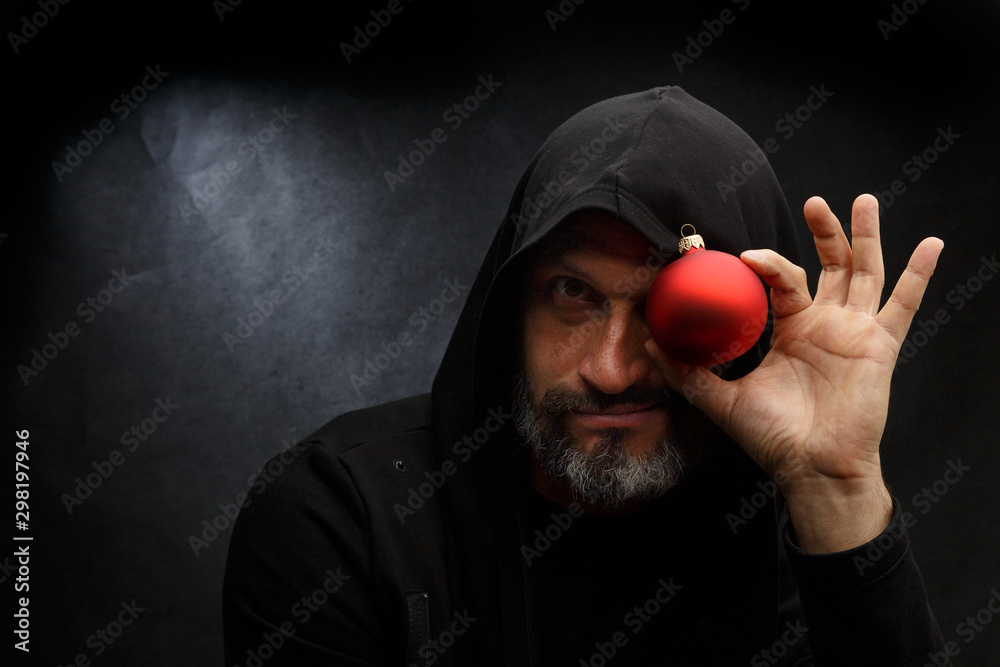 Wall mural Portrait of a bald man with a beard in a black hood witha red christmas ball on a dirty gray background. Young Santa concept.