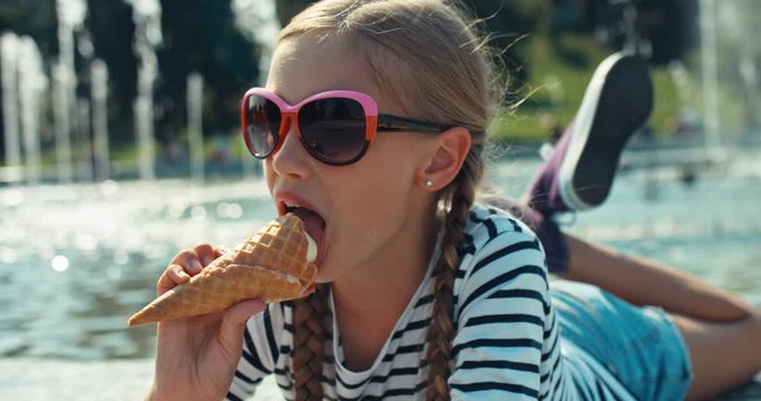 Close up portrait cheerful preteen girl in sunglasses eats ice cream and lying. Student has happy weekend in sunny day