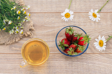 Glass cup of green tea, stawberries with white chamomile flowers on wooden background.