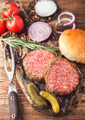 Fresh raw minced pepper beef burgers on vintage chopping board with buns onion and tomatoes on wooden background. Top view