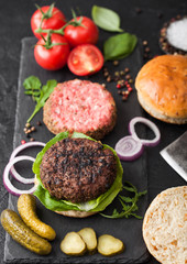 Fresh grilled and raw minced pepper beef burger on stone chopping board with buns onion and tomatoes on black background. Salty pickles and basil. Space for text