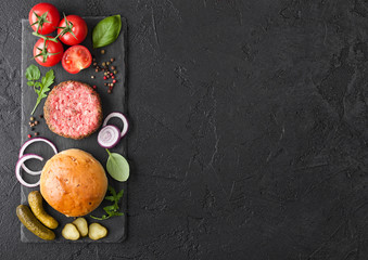 Fresh raw minced pepper beef burger on stone chopping board with buns onion and tomatoes on black background. Salty pickles and basil. Space for text