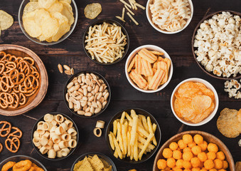 All classic potato snacks with peanuts, popcorn and onion rings and salted pretzels in bowl plates on wooden background. Twirls with sticks and potato chips and crisps with nachos and cheese balls.