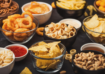 All classic potato snacks with peanuts, popcorn and onion rings and salted pretzels in bowl plates on black background. Twirls with sticks and potato chips and crisps with nachos and cheese balls.