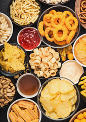 All classic potato snacks with peanuts, popcorn and onion rings and salted pretzels in bowl plates on black background. Twirls with sticks and potato chips and crisps with nachos and cheese balls.