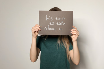 Young woman holding card with words IT'S TIME TO TALK ABOUT IT against light background