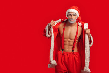 Christmas Freestyle. Young Santa Claus bare muscular upper body in hat standing isolated on red...