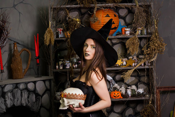 Fototapeta premium A young, beautiful girl in a green skirt, a black corset and a black hat holds a knife and a skull with a crown in her hands. Witch costume, Halloween, photo studio, model with clean skin.