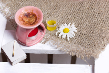 Aroma lamp stands on a wooden box. Nearby are oil in a cap and chamomile flowers.