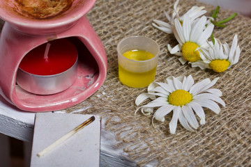 Aroma lamp stands on a wooden box. Nearby are oil in a cap and chamomile flowers.