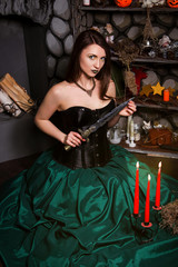 A young, beautiful girl in a green skirt and a black corset sits on the floor against a lighted candle with a knife in her hands. The witch conjures. Halloween.