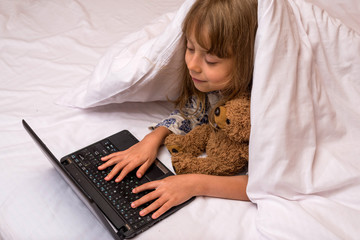 Fototapeta na wymiar Cute little girl using a laptop in bed under a blanket.
